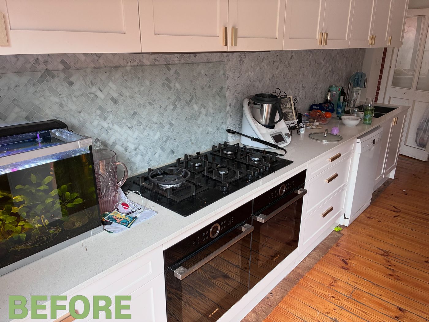 Before: a long white kitchen bench with bottles, a fruit bowl, a pot lid, cables and a jug crowded around the gas cooktop, next to a fish tank.